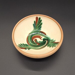 May include: A round ceramic bowl with a cream-colored interior and a light brown rim. The bowl features a green and red abstract design in the center. The design appears to be a stylized bird or floral motif.