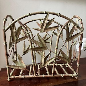 May include: A decorative metal magazine rack with a bamboo design. The rack features a light green and reddish-brown color scheme, with stylized bamboo stalks and leaves. The rack is designed to hold magazines or other reading materials.
