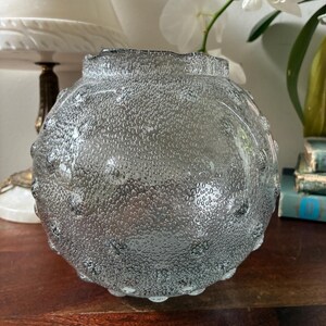 May include: A clear, round glass vase with a textured, bubble-like surface. The vase has a ruffled rim and sits on a wooden surface. The vase is a decorative item.