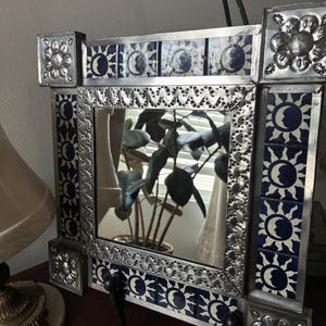May include: A square mirror featuring a frame decorated with blue and white tiles depicting sun and moon motifs. The frame is accented with embossed silver-toned metal borders and corner pieces featuring floral designs, supported by two metal legs.