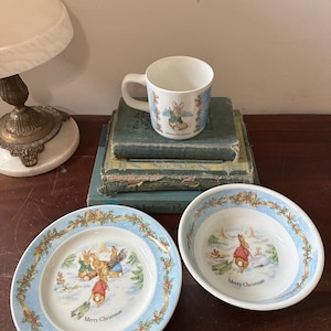 May include: A white ceramic mug, plate, and bowl set featuring a winter scene with rabbits and the text "Merry Christmas." The plate and bowl have a light blue border with holly and ribbon details. The mug is on a stack of books.