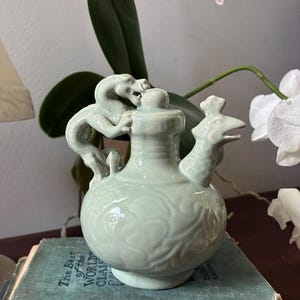 May include: A celadon-colored ceramic pitcher with a dragon handle and a bird-shaped spout. The pitcher has a bulbous body with a floral design and a small lid. The pitcher is sitting on a stack of vintage books.