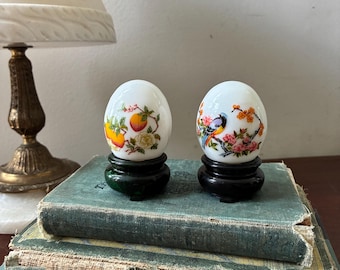 May include: Two white milk glass eggs decorated with colourful floral and fruit motifs, including oranges and a small bird perched on a branch. Each egg is displayed on a small, round, dark-coloured pedestal stand, creating a decorative set.