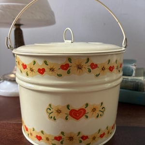 May include: Cream-colored metal container with a lid and a gold-colored handle. The container is decorated with a floral and heart pattern in red, yellow, and green. The container is round and has a lid.