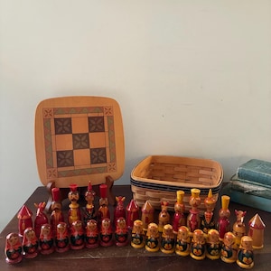 May include: A collection of hand-painted wooden chess pieces featuring stylised figures and towers in red, yellow, and black. The set includes a square wooden game board with a geometric pattern and a woven wooden basket for storage.
