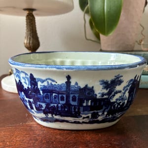 May include: A ceramic bowl featuring a white base with a detailed blue transferware pattern depicting a landscape with buildings, trees, and figures. The bowl has a smooth, rounded rim accented with a thin blue line and a fluted interior texture.