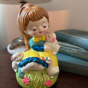 May include: A vintage ceramic figurine of a girl with brown hair, wearing a yellow dress and blue sleeves, holding a lamb. The base features a green grassy area with pink flowers, a white rabbit, and two white geese. The figurine has a glossy finish.