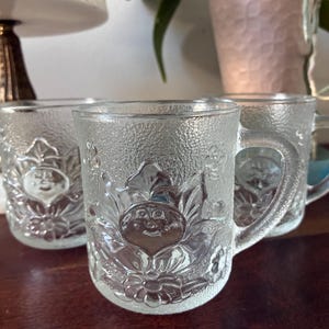 May include: A set of three clear glass mugs featuring a textured surface and a raised embossed design of a central flower with a smiling face surrounded by leaves and smaller floral accents, each with a sturdy handle and a smooth interior rim.