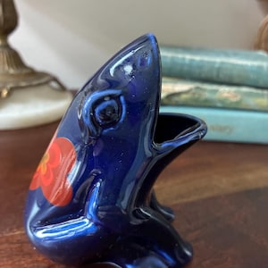 May include: A glossy dark blue ceramic frog figurine in a seated position with its mouth open. The frog features a stylized orange and red flower pattern on its back and side, with smooth, reflective surfaces and defined eye contours.