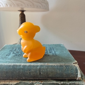May include: A small, orange-yellow rubber bunny figurine with a single visible eye, sitting atop a stack of antique books with teal covers. A white lamp with a decorative base is in the background.