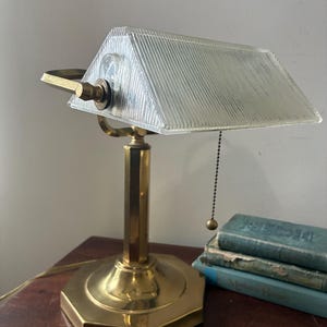 May include: A vintage brass desk lamp with a clear, ribbed glass shade. The lamp has a hexagonal base and a pull chain. The lamp is sitting on a wooden surface next to a stack of books.