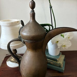 May include: A tall, weathered copper coffee pot featuring a bulbous base, a long curved spout, and a pointed lid with a decorative finial. The metal surface shows signs of oxidation and age, with a dark, muted patina across the entire exterior.