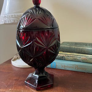 May include: A ruby red, vintage glass candy dish with a lid. The dish has a detailed, cut-glass pattern and sits on a pedestal base. The lid has a small, rounded finial. The dish is displayed on a wooden surface.