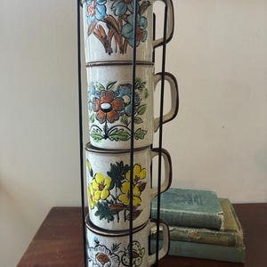 May include: A set of three ceramic mugs with a speckled, cream-colored glaze. Each mug features a different hand-painted design: a blue and green leaf, a yellow flower, and a red berry. The mugs have a simple, modern design.