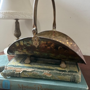 May include: An antique brass basket with a curved handle and a decorative painted interior. The basket rests on small claw feet and is displayed on top of two vintage books. The books are teal and green.