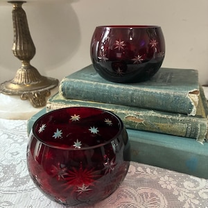 May include: Two round, deep red glass bowls featuring intricate, star-shaped cut patterns across their surfaces. The glass has a polished, reflective finish, and the star designs are etched into the exterior, creating a clear, contrasting texture against the dark red colour of the glass.