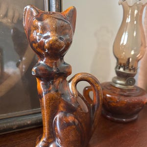 May include: A brown ceramic cat figurine with a glossy finish. The cat is sitting upright with its tail curled. The figurine has a bow tie detail and pointed ears. A vintage oil lamp is in the background.