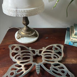May include: A silver-toned metal trivet shaped like a butterfly with an openwork design. The butterfly features symmetrical wing patterns with circular cutouts and a central body section with horizontal lines. The surface has a metallic finish.