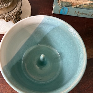 May include: A ceramic bowl with a glossy finish, featuring a two-tone design of white and teal. The interior of the bowl has a central, upward-pointing conical spike rising from the base, creating a functional shape for holding items.