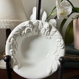 May include: A white ceramic bowl with embossed cherubs and roses, displayed on a dark metal stand. The bowl's rim features detailed relief designs of angels and floral elements. The bowl is supported by a dark metal stand.