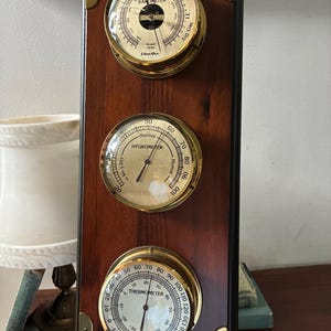 May include: A vertical wooden weather station featuring a brass-rimmed barometer, hygrometer, and thermometer. The barometer is labeled Ethan Allen and displays measurements in inches. The brass instruments are mounted on a dark wood panel with decorative brass corner accents.