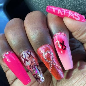 Op de afbeelding: Lange roze en rode acrylnagels met roze en witte lintontwerpen en strass. De nagels zijn versierd met het woord "TATAS" op één nagel.