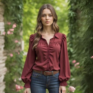 Puede incluir: Una mujer con una blusa de manga larga de color burdeos con un escote fruncido y un cinturón de cuero marrón. Lleva pantalones vaqueros azules y tiene el pelo largo y castaño.