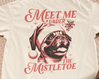 Könnte beinhalten: Cremefarbenes T-Shirt mit einem Mops, der einen Weihnachtsmannhut trägt. Die Grafik enthält den Text "Meet Me Under The Mistletoe" in festlicher roter Schrift. Das Design ist auf der Vorderseite des Shirts zentriert.
