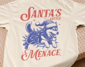 May include: Cream-colored t-shirt featuring a blue and white illustration of a dog wearing a Santa hat and Christmas lights. The text "Santa's Little Menace" is printed in red script.