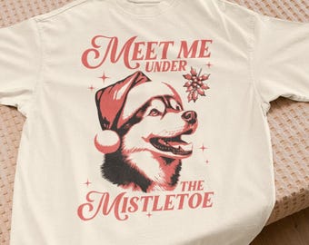 May include: Cream-colored t-shirt featuring a festive design. The graphic includes a dog wearing a Santa hat, with the text "Meet Me Under The Mistletoe" in red script. The design also includes a snowflake and small cross accents.
