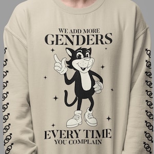 Pode incluir: Uma camisola bege com um gato de desenho animado preto e branco com um sorriso travesso. O texto "WE ADD MORE GENDERS EVERY TIME YOU COMPLAIN" está impresso na frente da camisola. As mangas da camisola têm um padrão repetido do símbolo de género.