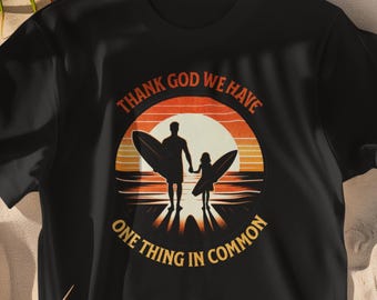 Può includere: Una t-shirt nera a maniche corte con una grafica circolare di un tramonto sull'oceano. Il design mostra le sagome di un adulto e di un bambino che tengono in mano delle tavole da surf, con il testo "THANK GOD WE HAVE ONE THING IN COMMON".