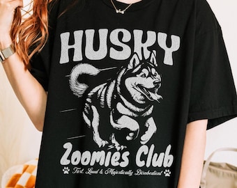 May include: Black t-shirt featuring a white graphic of a running husky dog and the text "HUSKY Zoomies Club". The text below the dog reads "Fast, Loud & Majestically Disobedient" with paw prints.