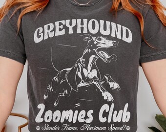 May include: Dark gray t-shirt featuring a white graphic of a running greyhound with the text "GREYHOUND Zoomies Club". The design includes the phrase "Slender Frame, Maximum Speed!" below the dog illustration.