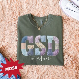 Puede incluir: Una camiseta verde musgo con las letras "GSD" en un diseño de patchwork y la palabra "mama" debajo. También se ven un collar dorado y zapatillas blancas.