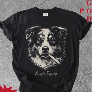 May include: Black t-shirt with a detailed illustration of an Australian Shepherd dog smoking a cigarette. The text "Aussie Issues" is written in cursive below the dog. The shirt has a crew neck design.