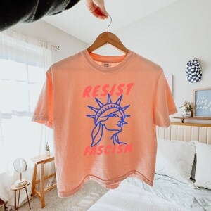 May include: Peach-colored t-shirt with the words "RESIST FASCISM" in pink and blue. The design features a blue outline of the Statue of Liberty. The shirt is hanging on a wooden hanger.