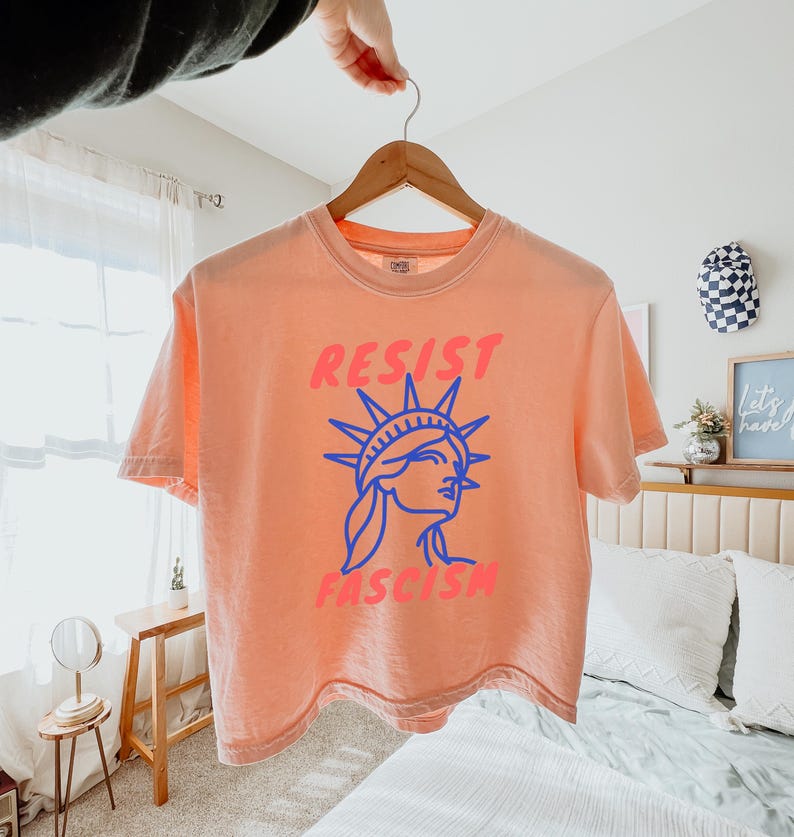 May include: Peach-colored t-shirt with the words "RESIST FASCISM" in pink and blue. The design features a blue outline of the Statue of Liberty. The shirt is hanging on a wooden hanger.