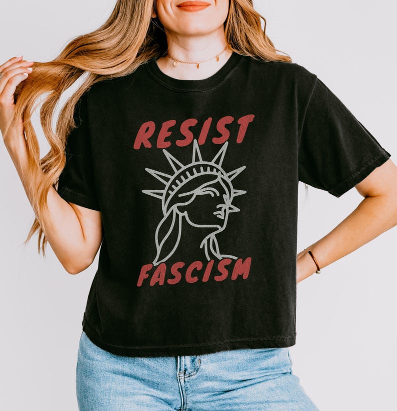May include: Black t-shirt with the words "RESIST FASCISM" in red and a white outline of the Statue of Liberty. The shirt is a casual style, suitable for everyday wear.