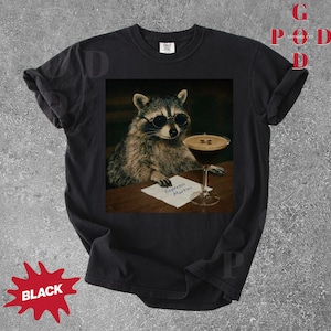 May include: Black t-shirt featuring a raccoon wearing sunglasses, holding an espresso martini. The raccoon is sitting at a table with a small sign that reads "Espresso Martini". The shirt has a distressed look.