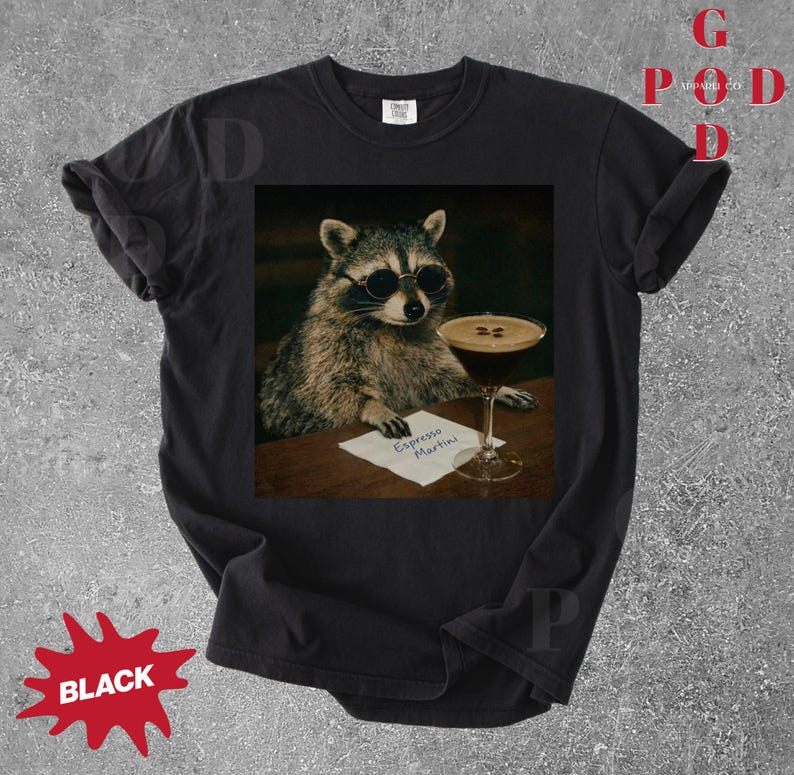 May include: Black t-shirt featuring a raccoon wearing sunglasses, holding an espresso martini. The raccoon is sitting at a table with a small sign that reads "Espresso Martini". The shirt has a distressed look.