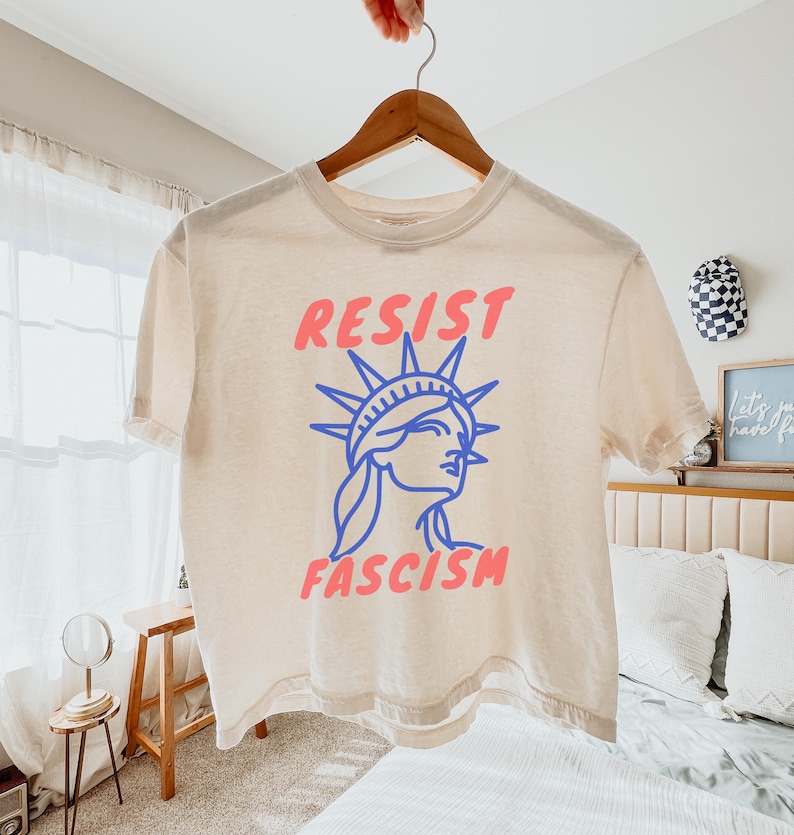 May include: Cream-colored t-shirt with the words "RESIST FASCISM" in pink and blue. The design features a blue outline of the Statue of Liberty. The shirt is hanging on a wooden hanger.