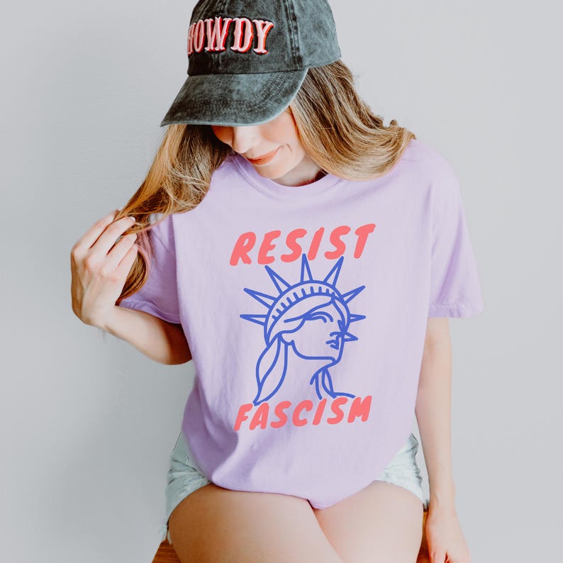 May include: A light purple t-shirt with the words "RESIST FASCISM" and a blue graphic of the Statue of Liberty. The person is wearing a black cap with the word "HOWDY" in pink.