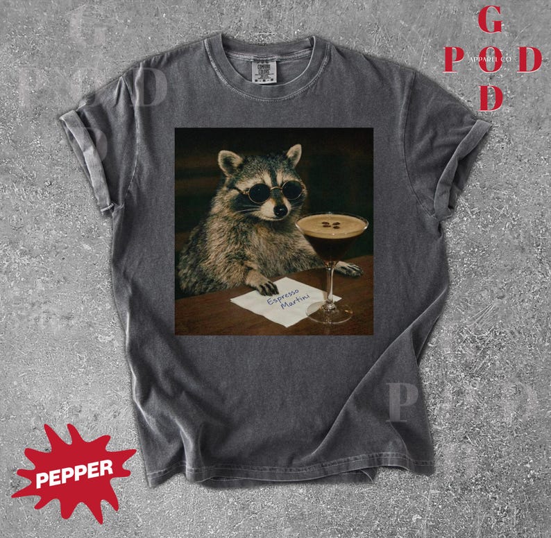 May include: Dark gray t-shirt featuring a raccoon wearing sunglasses, seated at a table with an espresso martini. The raccoon is holding a small paper with the words "Espresso Martini" written on it. The shirt has rolled-up sleeves.