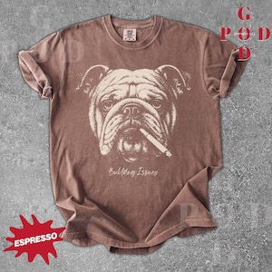 May include: Espresso-colored t-shirt featuring a detailed illustration of a bulldog with a cigarette. The graphic is in a cream color, and the text "Bulldog Issues" is below the dog's face. The shirt has rolled-up sleeves.