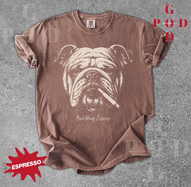 May include: Espresso-colored t-shirt featuring a detailed illustration of a bulldog with a cigarette. The graphic is in a cream color, and the text "Bulldog Issues" is below the dog's face. The shirt has rolled-up sleeves.