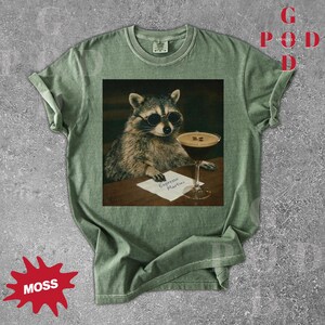 May include: Moss green t-shirt featuring a raccoon wearing sunglasses, holding a pen, and sitting next to an espresso martini. The text "Espresso Martini" is on a small paper. The shirt has a vintage look.