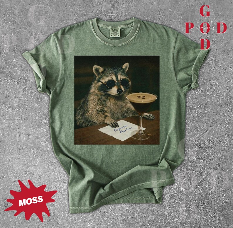 May include: Moss green t-shirt featuring a raccoon wearing sunglasses, holding a pen, and sitting next to an espresso martini. The text "Espresso Martini" is on a small paper. The shirt has a vintage look.