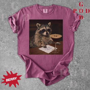 May include: Berry-colored t-shirt featuring a raccoon wearing sunglasses, holding an espresso martini. The raccoon is depicted in a vintage style, with the words "Espresso Martini" on a small paper. The shirt has a crew neck and short sleeves.