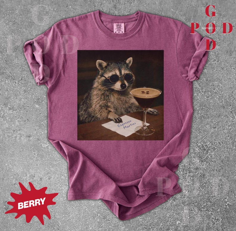 May include: Berry-colored t-shirt featuring a raccoon wearing sunglasses, holding an espresso martini. The raccoon is depicted in a vintage style, with the words "Espresso Martini" on a small paper. The shirt has a crew neck and short sleeves.
